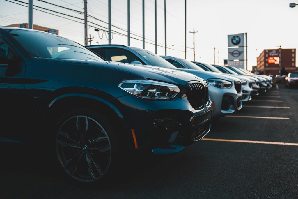 Một hàng xe BMW nhiều màu đỗ nối nhau trước bảng hiệu đại lý lúc chiều muộn