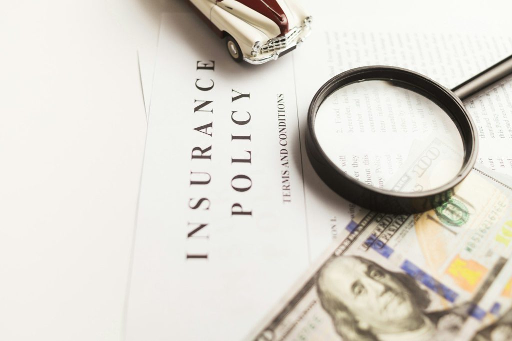 Tờ giấy in chữ Insurance Policy, chiếc kính lúp, mô hình ô tô cổ màu kem và tờ tiền đô la đặt trên bàn trắng