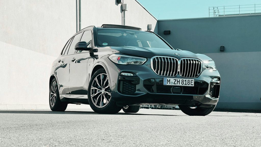 BMW X5 màu xám đậm với lưới tản nhiệt hình quả thận lớn, đỗ trên sân bê tông cạnh bức tường trắng
